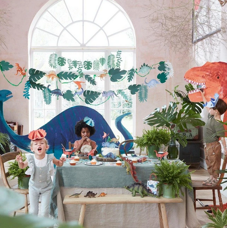 Meri Meri dinosaur party collection image of children at a party table with large dinosaur cut out behind them and the dinosaur garland hanging above the party table.