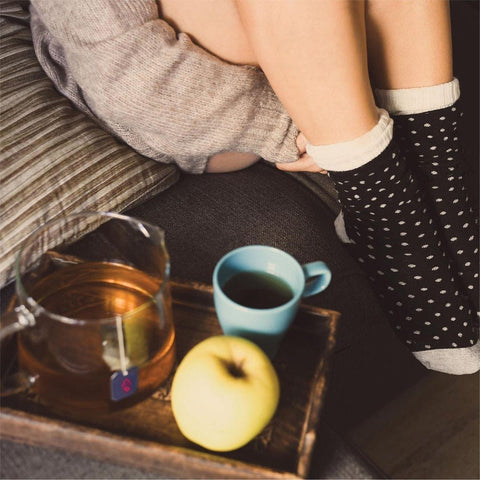 Shot of a girl's legs with black socks, and a tray showing in the foreground with a tea cup, a tea pot containing a Zest tea bag, and an apple.