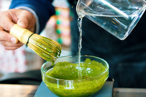 Water being poured into a bowl of matcha tea that's being mixed by a bambo whisk.