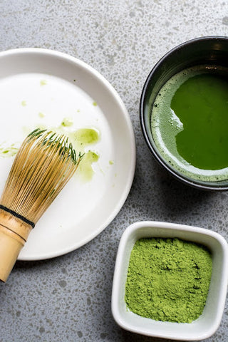 Bowl with matcha, a bamboo whisk, and a cup of matcha powder on a table