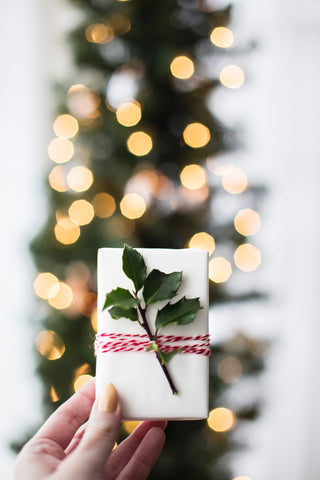 A gift wrapped with plain white paper, candy cane inspired string and a a Christmas tree stands in the background
