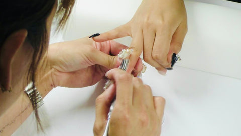 Close-up of a nail artist carefully placing a rhinestone onto a nail art pattern on a student's finger. Her nails are painted black.