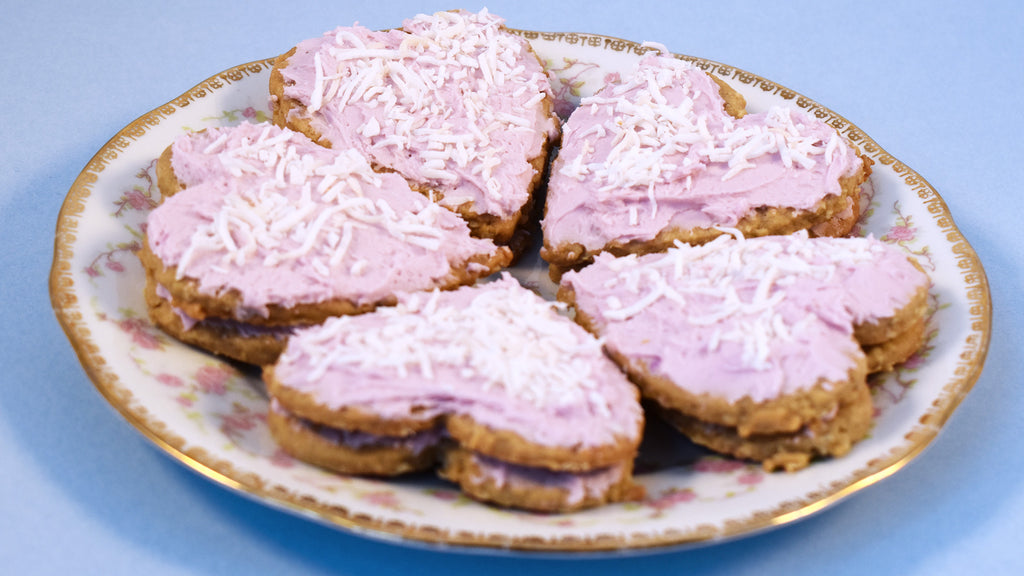 A plate of five lavender valentine sandwich cookies.  Each cookie sandwich is made up of two oatmeal cookies, with pink frosting between and on top. There is shredded coconut sprinkled atop  each one. They rest on a china plate with a rose pattern and gold edges.