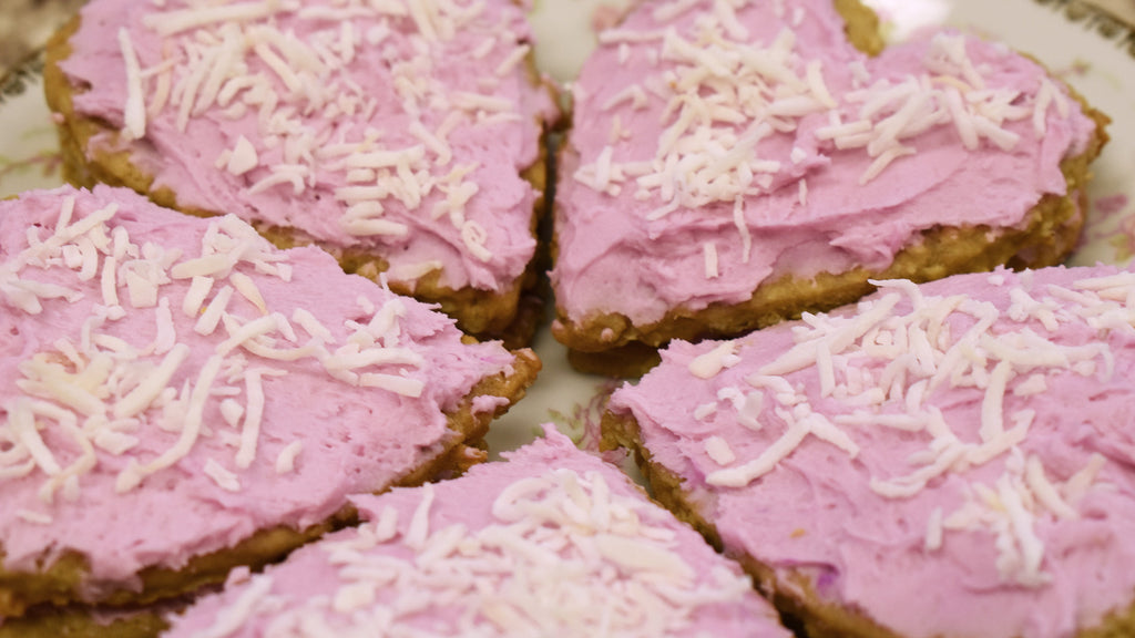 A close up photo of Lavender Valentine Sandwich Cookies - two oatmeal cookies stacked on top of one another with pink frosting between and on top. A sprinkling of shredded coconut sits atop each one. Pictured are five cookies on a gold-edged china plate with a rose pattern.