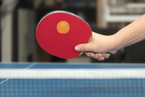 Close up of person holding ping pong paddle and orange ping pong ball on blue table.