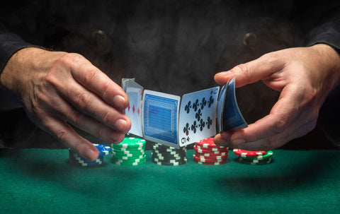 Close-up of the hands of a dealer shuffling poker cards in a smoky club on a green poker table with chips. 