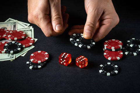 Man holding two black poker chips on a black poker table with poker chips, money, and dice. 