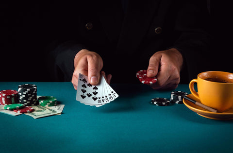 Man in black suit holding five poker cards on a green poker table with chips and money and a yellow coffee cup on a yellow dish with a cigarette.