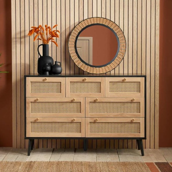 Rattan 7 drawer chest with black wood frame. Wood panelling with a round mirror on the wall in the background. Black vases on top of the chest.