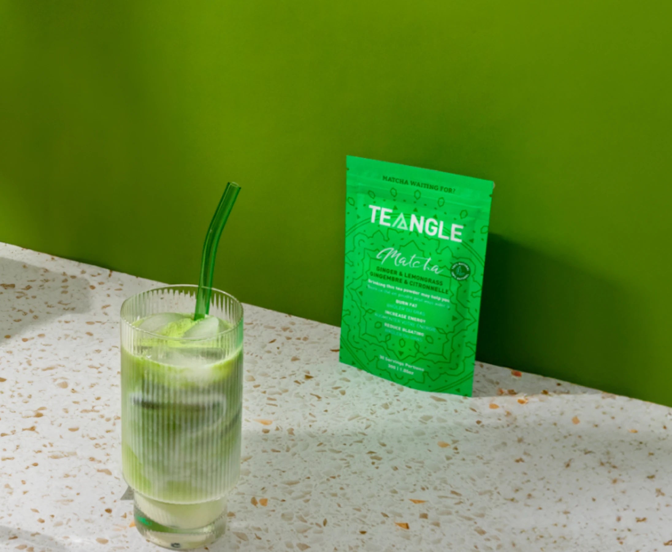 Iced matcha drink with a green straw beside a packet of matcha powder on a speckled countertop.