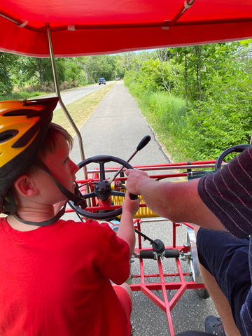 young boy looking at man's arm while steering 4 person bike at Presque Isle State Park with a red awning over top of them 