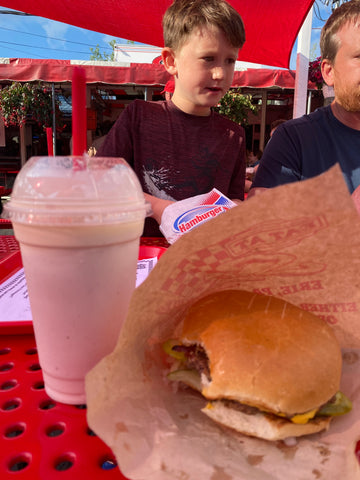 cheeseburger wrapped in kraft paper with a bite taken and a milkshake with whipped cream and a red straw, young boy in background 