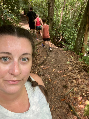 selfie photo of woman in white tank top with family hiking ahead of her 