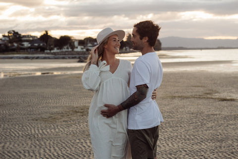 Maternity photoshoot of couple at beach, women wearing a off white maternity dress
