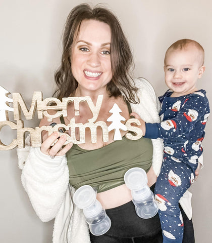 Woman holding a young toddler while using a breast pump. She is also holding a wooden sign that says "Merry Christmas".