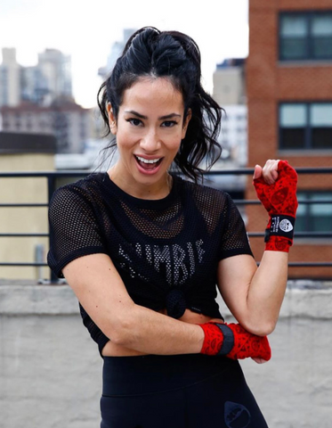 Mona Lavinia smiling in a black Rumble Boxing shirt and red boxing practice gloves.