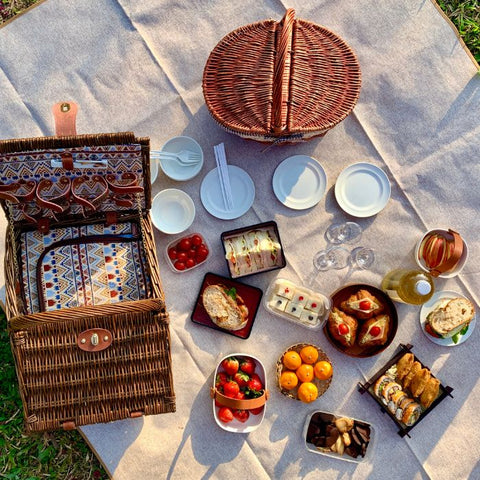 Picnic baskets, fruits, sandwiches, and plates spread on a blanket in grass