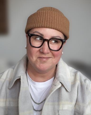 A photo of author Kelly Small. They're wearing a white t-shirt with a button up over top, a brown beanie hat, and are wearing black thick framed glasses.