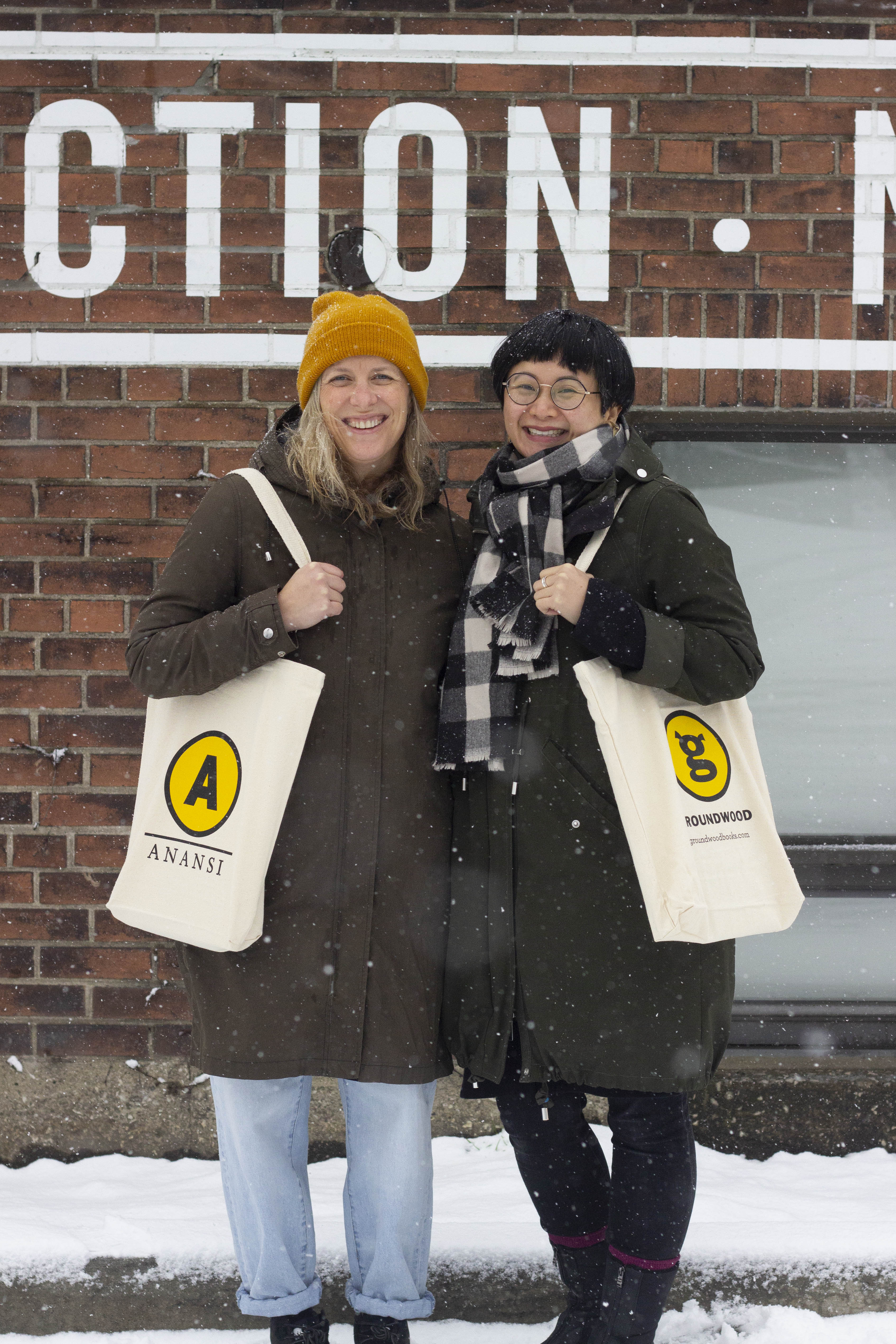 A picture of Leigh Nash and Karen Li. Leigh holds an Anansi tote bag and Karen holds a Groundwood tote bag. They are standing in front of a brick wall in the snow. 