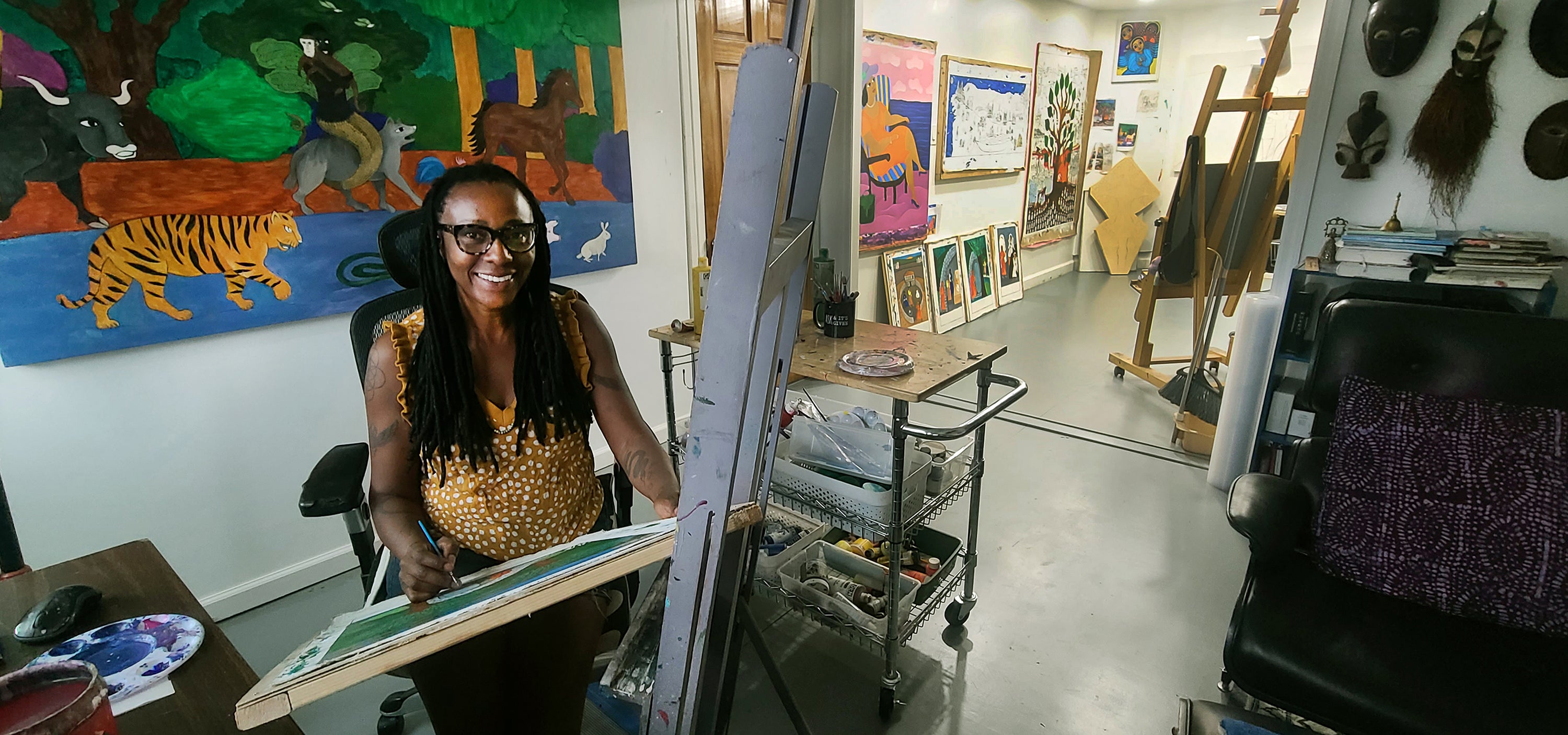 A photo of author and illustrator Laura James sitting at her easel in her arts studio. There are large scale paintings on the wall behind her and other painting supplies/ apparatus.