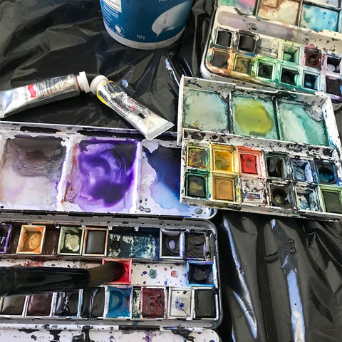 A photo of watercolour paint trays resting on a black vinyl table cloth. A yogurt for water container sits next to the open trays.