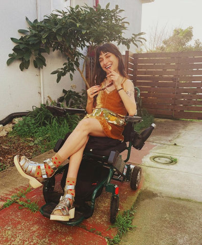 A photo of Maria Sweeney in her power chair outside in the sun. She has taken off her sunglasses and is smiling at the camera.