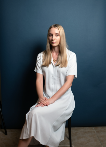 A photo of Cassidy McFadzean in a white dress sitting on a stool, looking at the camera. Viewers see her entire seated figure. Photo by Tony Tulathimutte.