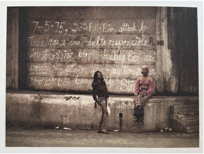A photo of two people posing for the camera at a loading dock. The dock door behind them is graffitied. One of the people has long black, loosely curled hair, and is wearing denim shorts and a dark long-sleeve jacket. They have a medium-dark skintone. The other person is wearing a pink headscarf and a matching pink crop top with long balloon sleeves and an off-the-shoulder neckline, as well as long pants or a long skirt that sparkles. Around the pair garbage is strewn, and the concrete is water-stained..
