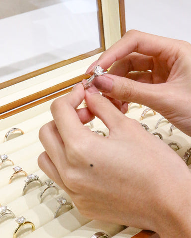 Slender hand holding an engagement ring at the tip of fingers. The holder has light pink nails and the engagement ring features a solitaire design with a round colorless diamond at least 1.5 carats. It is being held on top of a jewelry box with a variety of ring designsl.