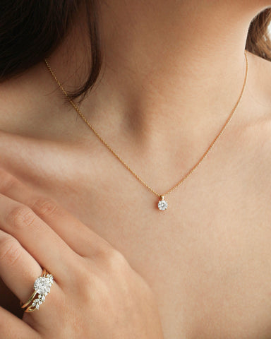 A tan-skinned woman with black medium length hair wears a yellow gold solitaire necklace with a round diamond that falls in between her collarbones. Her hand rests below her right collarbone, with the ring finger adorned with a round solitaire diamond engagement ring stacked flush with the Fern wedding band (a design inspired by nature characterized by round and marquise diamonds).