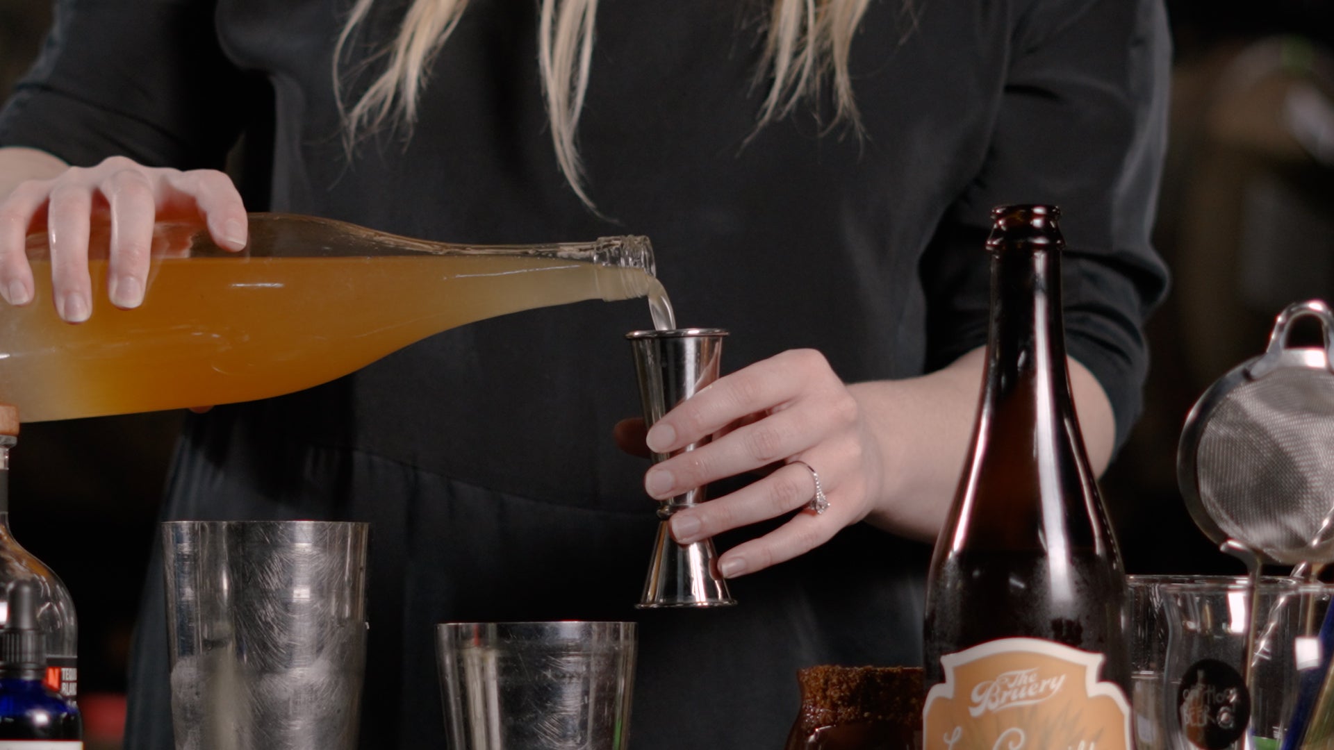 Close up of pouring a bottle of orange liquid (the shrub from the La Doña cocktail) into a jigger, surrounded by other ingredients.