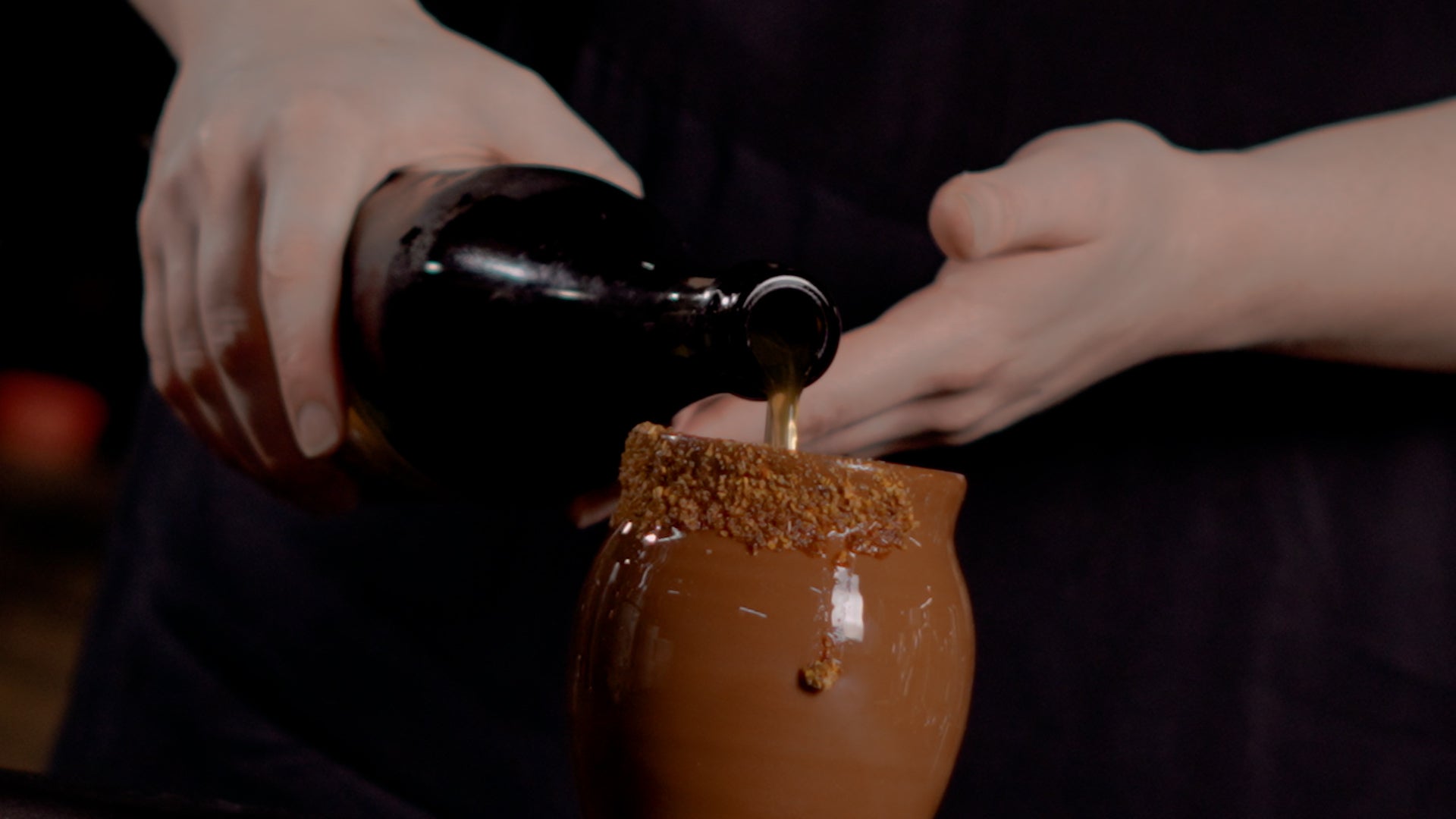 Close up of pouring a bottle of La Capilla into a brown mug with a tajin rim