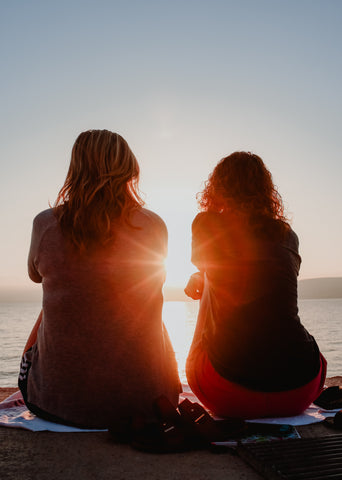 zwei Frauen sitzen auf einer Decke am Wasser und schauen in den Sonnenuntergang