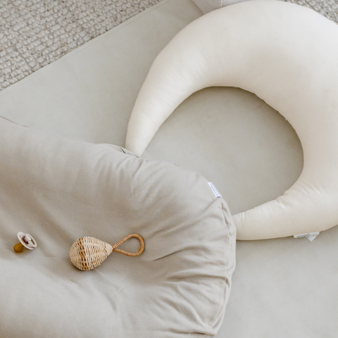 A breastfeeding support pillow and baby support cushion sit on the floor as potential items to purchase for a baby gift registry. 