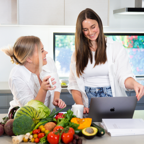 co founders of mother's mylk and authors of nourished postpartum doula and naturopath in kitchen with fresh fruit around them smiling at the work they do for pregnant and postpartum mothers