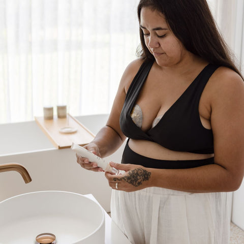 A new mum stands in the bathroom with breast care products. She is wearing a maternity bra and postpartum briefs as well as hot and cold compresses for the breasts. In her hand is a breast care balm.