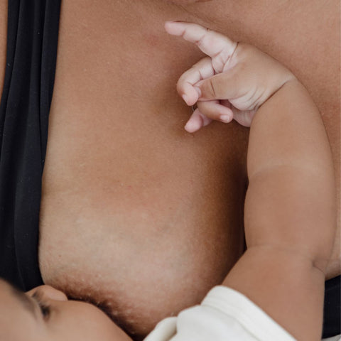 A close up image of a baby breastfeeding and establishing breastfeeding with its mother. She is gazing down at the baby while the baby looks up and reaches up towards her. It’s a sweet bonding moment between mum and baby.