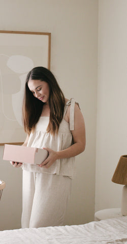 A pregnant woman in maternity clothes for the first trimester stands and looks at a pregnancy gift box.