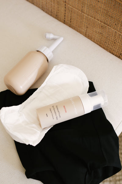 A pair of maternity briefs and postpartum briefs sit on a bench. On top of them is a maternity pad, perineal wash bottle, and perineal foam for postpartum pain relief. Together they make up a recovery kit after labour.