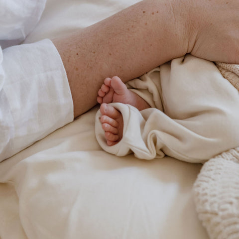 A close up image of little baby toes poking out from an oversized onesie. A mum’s hand lays next to the baby’s feet. It’s a precious image of an intimate moment between mum and newborn.
