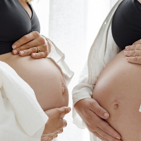 Two women stand facing toward one another with their bare pregnant bellies nearly touching. They are embracing the preparation of birth together.