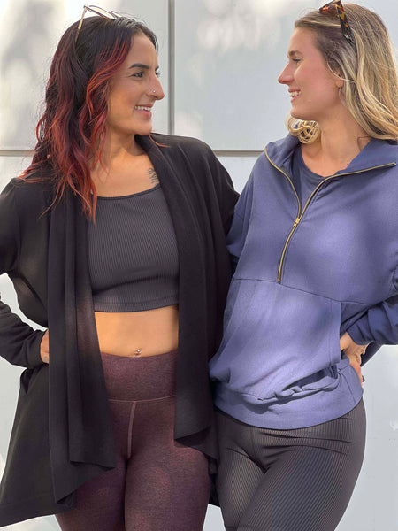 Two girls wear clothing from Vancouver women's clothing brand, DAUB. The woman on the left wears a ribbed sports bra with a fleece jacket and leggings. The other girl wears a half zip crewneck sweatshirt in navy blue.