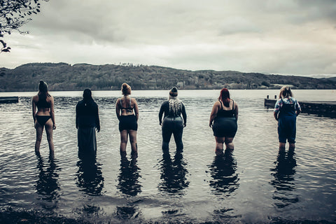 A row of women all standing in a line at the water's edge looking away from the camera. 
