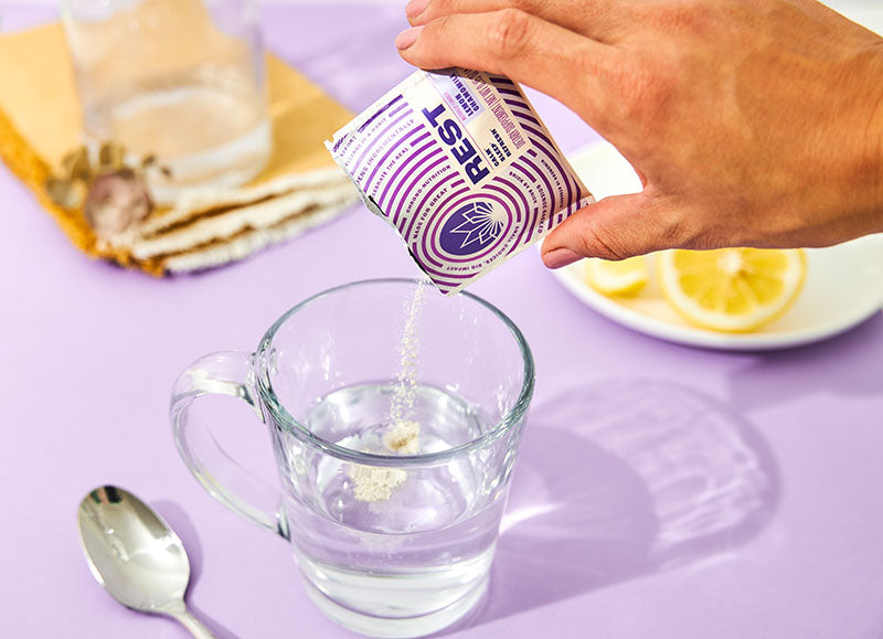 A person pouring a REST supplement from MANTRA Labs into a glass of water against a purple background