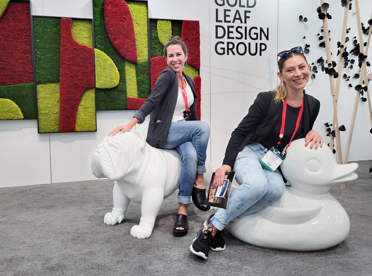 Two women sitting on large white fiberglass scuptures (a duck and a bulldog) at a Gold Leaf Design Group tradeshow display.