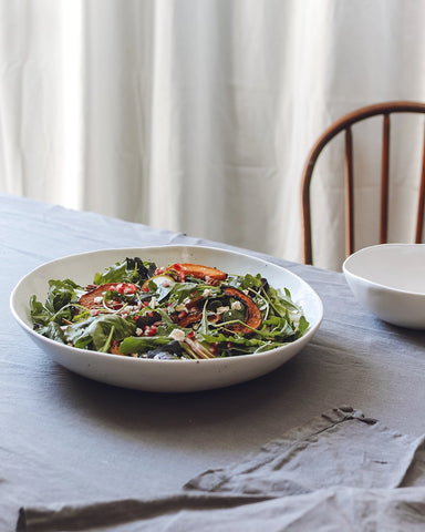 Fresh arugula and mixed greens salad with roasted carrots and pomegranate seeds in a handmade white ceramic bowl, presented on a grey tablecloth against a backdrop of sheer white curtains.