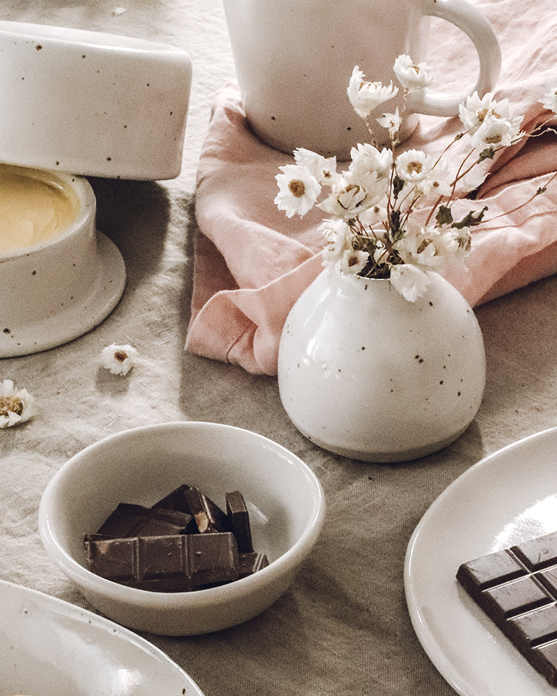 A serene Easter setting with a bowl of dark chocolate chunks and a simple ceramic vase holding delicate white flowers, complemented by soft pink linen, creating a harmonious blend of natural tones and textures.