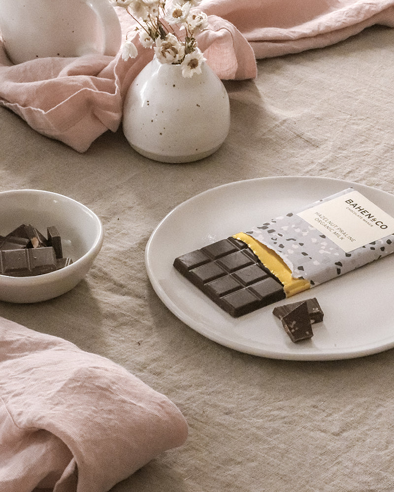 Easter indulgence with a stylishly packaged chocolate bar partially unwrapped on a ceramic plate, surrounded by scattered chocolate pieces, white flowers in a vase, and a pink linen napkin for a touch of spring elegance.
