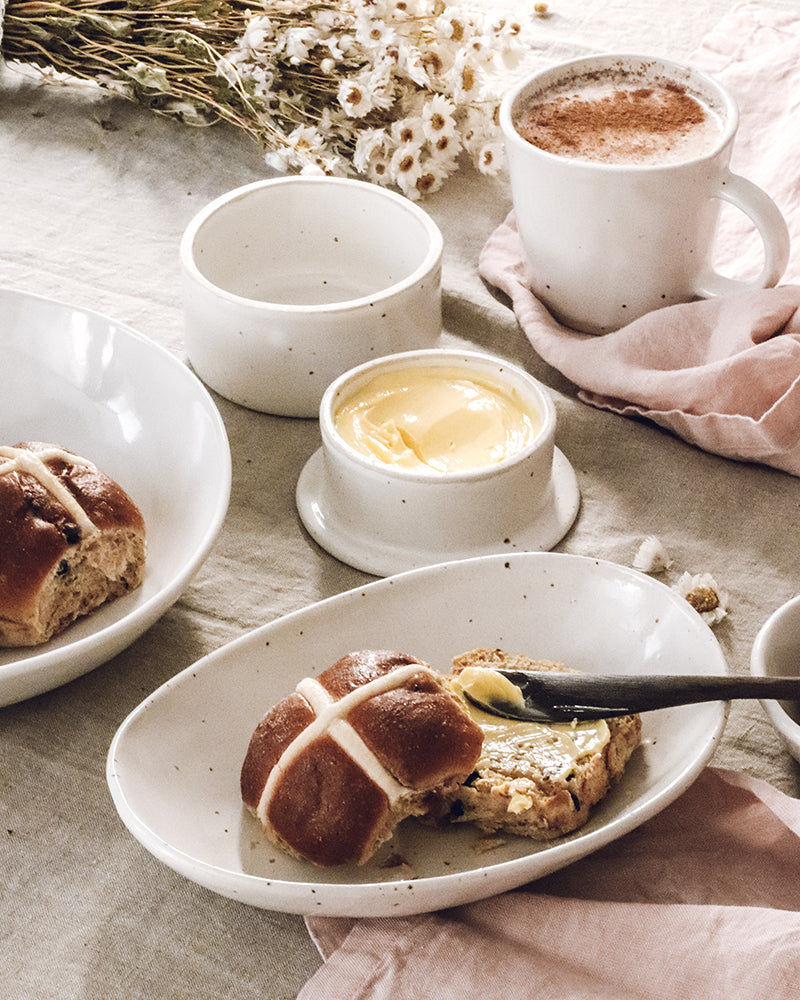 Close-up of a handmade ceramic pot of golden butter and a plate with a hot cross bun, with a textured spoon resting beside it, invoking a sense of warmth and homemade Easter comfort