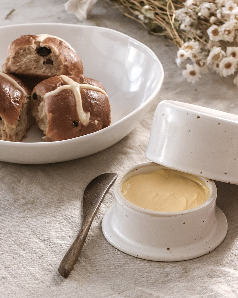 Close-up of a handmade ceramic pot of golden butter and a plate with a hot cross bun, with a textured spoon resting beside it, invoking a sense of warmth and homemade Easter comfort.